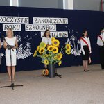 Kobiety prowadzące uroczystość zakończenia roku szkolnego 2024/2025 stoją przy mikrofonie. Ob-ok nich, przy stole, widoczna dekoracja z dużymi sztucznymi słonecznikami. W tle stoją uczniowie w białych koszulach i czarnych spódnicach, trzymający sztandar Polski. Na sali gimnastycznej odbywa się uroczysta ceremonia, a w tle widoczny jest napis „Uroczyste zakończenie roku szkolnego 2024/2025”.