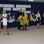 Uroczystość zakończenia roku szkolnego 2024/2025. Na pierwszym planie stoją kobiety prowadzące i ksiądz, który wygłasza przemówienie. W tle widoczny poczet sztandarowy, trzymający sztandar Polski, a także dekoracja z napisem „Uroczyste zakończenie roku szkolnego 2024/2025” oraz sztucz-nymi słonecznikami w donicy. Uczniowie w odświętnych strojach są ustawieni na sali gimnastycznej, a ceremonia ma uroczysty charakter.