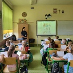 Policjantka w mundurze prowadzi prelekcję dla uczniów pierwszej klasy, siedzących w szkolnych ławkach. Dzieci uważnie słuchają, a w tle widoczna jest tablica z elementami prezentacji. Spotkanie odbywa się w jasnej, kolorowo udekorowanej sali lekcyjnej.