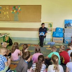 Policjantka w mundurze siedzi na krzesełku przed grupą dzieci zgromadzonych na dywanie i pokazuje znak przejścia dla pieszych. Uczniowie z zaciekawieniem słuchają i obserwują prezentowany znak. Zajęcia odbywają się w kolorowej sali lekcyjnej, w przyjaznej i edukacyjnej atmosferze.
