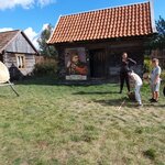 Na zdjęciu widać trójkę chłopców, który stoją w rzędzie i przygotowują się do rzutu ziemniakiem do tarczy. Oprócz nich jest też pani, która jest ubrana cała na czarno. Pierwszy chłopiec pochyla się do koszyka i bierze ziemniaka i patrzy w kierunku tarczy stojącej z lewej strony zdjęcia. Dwóch pozostałych chłopców patrzy się w kierunku aparatu. Widać także dwa drewniane domki. Jeden z nich ma plakat z królem, drugi drewnianą okiennicę.