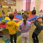 Dzieci stoją w kręgu trzymając chustę animacyjną. Na chuście są dwie piłki_ które dzieci p.jpg