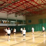 Uczniowie w strojach sportowych biorą udział w zajęciach z unihokeja na sali gimnastycznej. Każdy trzyma kij do gry i jest aktywnie zaangażowany w ćwiczenia. W tle widoczne są zielone kotary oddzielające sektory sali, kosze do koszykówki oraz ławki. Pomieszczenie jest dobrze oświetlone światłem dziennym.