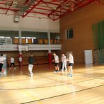 Grupa uczniów w strojach sportowych rozstawiona jest na sali gimnastycznej. Jeden uczeń kopie piłkę. W tle widać bramkę, kosz, trybuny, baner z napisem 