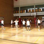 Grupa uczniów w strojach sportowych stoi na sali gimnastycznej w trakcie zajęć wychowania fizycznego. Część dzieci trzyma piłkę, inni stoją w rzędach. W tle widać bramkę, kosz, nauczyciela oraz uczniów oczekujących przy ścianie sali. Pomieszczenie jest dobrze oświetlone światłem dziennym.
