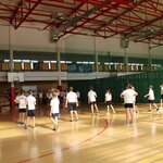Uczniowie w białych koszulkach i ciemnych spodenkach uczestniczą w zajęciach wychowania fizycznego na sali gimnastycznej. Dzieci stoją w grupach, słuchając nauczyciela. W tle widoczna siatka oddzielająca sektory, banery sportowe oraz trybuny z ławkami.