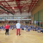 Duza grupa uczniów siedzi na podłodze w hali _ z przodu widoczni dwoje ludzi stojący tyłem.jpg