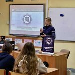 Na zdjęciu widzimy policjantkę stojącą przed grupą uczniów w sali lekcyjnej. Za nią znajdują się tablice. Na jednej z nich widoczny jest napis 