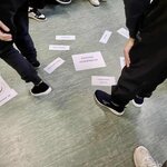  Na zdjęciu widzimy grupę dzieci lub młodzieży stojącą wokół kartek rozłożonych na podłodze. Na kartkach znajdują się hasła, które stanowią część zajęć edukacyjnych dotyczących przemocy rówieśniczej.