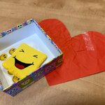 Na zdjęciu widzimy kolorowe pudełko. W środku znajduje się miękka, żółta poduszka w kształcie kwadratu z wyszytą uśmiechniętą buźką oraz kilka naklejek z podobnymi uśmiechniętymi emotkami. Pudełko stoi na drewnianym blacie, a obok niego leży duże, czerwone, papierowe serce – lekko pogniecione i przyklejone taśmą klejącą.
