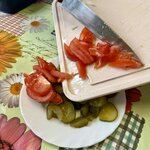 Na zdjęciu widać osobę przekładającą pokrojone pomidory z deski do krojenia na talerz, na którym leżą również plasterki ogórków.