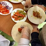 Na zdjęciu widać osoby przygotowujące tortille. Na stole ułożone są składniki: pokrojone warzywa, mięso, sałata i sosy.