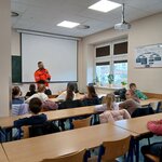 Ratownik opowiada o numerach alarmoawych w sali szkoleniowej. Uczniowie siedzą w ławkach...jpg