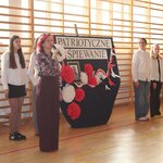Pani dyrektor stoi_ w tle widać troje uczniów  oraz tablicę z napisem Patriotyczne Śpiewanie z białymi i czerwonymi kwiatami.jpg