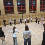 Zdjęcie przedstawia salę gimnastyczną w szkole, w której odbywają się zajęcia sportowe. Na środku sali widać dużą grupę uczniów ustawionych w szerokim okręgu. Uczniowie stoją na drewnianej podłodze boiska, na której widoczne są linie wyznaczające pola gry. W tle znajduje się ściana z drewnianymi drabinkami gimnastycznymi oraz duże okna z czerwonymi ramami. W kilku miejscach na podłodze ustawione są żółte pachołki, wyznaczające strefy ćwiczeń. Oświetlenie jest jasne, a cała scena wygląda na część zajęć ruchowych.