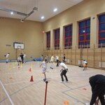 Zdjęcie przedstawia salę gimnastyczną w szkole, w której odbywają się zajęcia sportowe. Na drewnianej podłodze boiska ustawione są kolorowe pachołki i znaczniki, wyznaczające strefy ćwiczeń. Widać grupę uczniów aktywnie uczestniczących w zabawie z piłkami – część osób rzuca lub odbija piłki, a inne przygotowują się do kolejnych ćwiczeń. W tle znajduje się ściana z dużymi oknami w czerwonych ramach oraz drewnianymi drabinkami gimnastycznymi. Na lewej stronie widoczny jest kosz do koszykówki. Oświetlenie jest jasne, a atmosfera dynamiczna, typowa dla zajęć ruchowych.