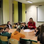 Zdjęcie przedstawia salę szkolną z dużym stołem, wokół którego siedzi grupa uczniów. Na stole leżą kartki papieru i przybory, odbywają się warsztaty . Uczniowie siedzą na zielonych krzesłach, a w tle widać okna z pionowymi żółtymi i zielonymi żaluzjami. Na ścianie znajduje się biały ekran do projekcji, a nad nim widoczne są symbole państwowe. Oświetlenie jest jasne, typowe dla sali lekcyjnej.