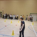 Zdjęcie przedstawia salę gimnastyczną w szkole. Na pierwszym planie znajduje się uczeń w ciemnym stroju sportowym, który porusza się w kierunku aparatu. Na podłodze ustawione są żółte pachołki i czerwone znaczniki, tworzące tor przeszkód. W tle, przy ścianie z jasnych cegieł, stoi grupa uczniów w różnorodnych strojach, czekających na swoją kolej. Po prawej stronie widoczny jest fragment kosza do koszykówki oraz zwinięta mata gimnastyczna. Całość odbywa się w jasnym, dobrze oświetlonym pomieszczeniu sportowym.