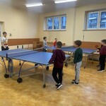Grupa kilku chłopców gra w tenisa stołowego w jasnej sali szkolnej. W pomieszczeniu stoją dwa stoły do ping-ponga ustawione obok siebie, a dzieci trzymają rakietki i aktywnie uczestniczą w grze