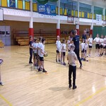 „Grupa dzieci w szkolnej sali gimnastycznej uczestniczy w zajęciach sportowych. Uczniowie stoją w kilku rzędach, trzymając piłki do rugby, a dwóch dorosłych prowadzących znajduje się pośrodku. W tle widać kosze do koszykówki, zielone zasłony oddzielające sektor sali oraz ławki gimnastyczne przy ścianach.”
