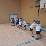 Zdjęcie przedstawia grupę dzieci w Sali gimnastycznej. Dzieci stoją  w czterech małych grupkach. Wykonują ćwiczenie polegające na podtrzymywaniu piłki głową. Piłki są niebieskie i pomarańczowe