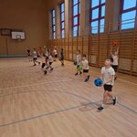 Zdjęcie przedstawia grupę dzieci w sali gimnastycznej podczas zajęć sportowych. Dzieci stoją w kilku rzędach i trzymają kolorowe piłki . Wykonują ćwiczenie polegające na podnoszeniu piłek przed sobą. Atmosfera jest dynamiczna, jak podczas typowej lekcji wychowania fizycznego.