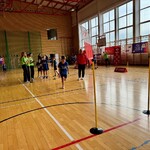 „W dużej sali gimnastycznej odbywają się zawody sportowe dla dzieci. Chłopiec w niebieskiej koszulce biegnie między dwoma żółtymi tyczkami ustawionymi na parkiecie. Po lewej stronie stoi grupa dzieci oraz osób w zielonych koszulkach, które mierzą i notują wyniki. W tle widać banery, sprzęt sportowy oraz osoby obserwujące wydarzenie.”