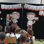 Dziewczynka z mikrofonem stoi przed publicznością_ w tle tablica z dekoracją.jpeg
