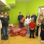 
Grupa osób stoi w jasnym pomieszczeniu o zielonych ścianach i żółtej podłodze. W centrum znajdują się duże stosy kolorowo zapakowanych prezentów w czerwonym papierze z różnymi wzorami. Kilka osób trzyma paczki w rękach, a reszta stoi obok, tworząc półokrąg wokół podarunków. W tle widoczne są drzwi prowadzące do innego pomieszczenia oraz fragment metalowej siatki po lewej stronie. Są to przygotowania do świątecznej akcji Szlachetna Paczka.