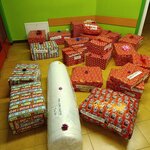 Na zdjęciu widać stos dużych paczek zapakowanych w kolorowy papier prezentowy, ułożonych na podłodze w rogu pomieszczenia o jasnozielonych ścianach i żółtej podłodze. Większość paczek jest owinięta w czerwony papier z białymi wzorami, a niektóre w papier z napisem „HO HO HO” lub z motywem świątecznym. Na kilku paczkach znajdują się ozdobne kokardki w kolorze fioletowym i różowym. Wśród prezentów leży także jedna duża, cylindryczna paczka owinięta w biały materiał. Wszystkie paczki mają przyklejone etykiety z kodami i numerami Szlachetnej Paczki.