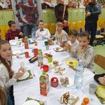 Na głównym planie widać ośmioro dzieci, którzy siedzą przy stole. Czwórka z nich patrzy na robione im zdjęcie, a pozostali patrzą się przed siebie. Jedna dziewczynka z lewej strony, w białej bluzce je pierniczka. Za nią siedzi chłopiec w czarno-czerwonej koszuli, który patrzy się na stół, dziewcyznka w aureoli, chłopiec w czerwono-granatowej koszuli, który chce wziąć coś ze stołu. Na wprost, kolejno siedzi chłopiec w białej koszuli i ciemnej muszce, który się uśmiecha. Następnie siedzi chłopiec w iałym T-shircie i futerku, który coś przeżuwa, chłopiec w białym T-shircie i chłopiec z kucykiem w bluzce z długim rękawem z jakimiś napisami, który trzyma paluszki w dłoni. Na stole stoją przekąski, lampiony, zastawa papierowa i napoje.