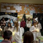 Na zdjęciu widać osiem dziewczynek przebranych za aniołki, które tańczą w dwóch kołach z zapalonymi lampionami. W tle widać siedmioro dzieci, którzy stoją i są zwróceni do przodu. Za nimi stoją dwa granatowe parawany z dekoracją świąteczną oraz widzą dwie tablice korkowe z dekoracją zimową.