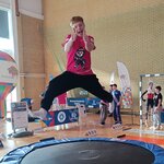 W sali gimnastycznej dziecko w różowej koszulce wykonuje skok na małej trampolinie, unosząc nogi szeroko na boki. W tle widoczne są stanowiska sportowe z tablicami informacyjnymi, kolorowymi banerami oraz grupa obserwujących osób. Na podłodze stoją elementy wykorzystywane do prezentacji zimowych aktywności sportowych.