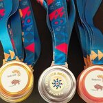 Na zdjęciu widoczne są trzy grupy medali sportowych: złote, srebrne i brązowe. Każdy medal zawieszony jest na szerokiej, niebieskiej wstędze z kolorowymi geometrycznymi wzorami. Na medalach znajdują się logotypy oraz napisy związane z wydarzeniem sportowym organizowanym w Białymstoku. 