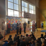 W sali gimnastycznej odbywa się wydarzenie sportowe. Na podłodze siedzi grupa dzieci zwróconych w stronę sceny. Przed nimi stoją prowadzący oraz osoby przemawiające. W tle widoczna jest duża dekoracyjna ścianka z kolorowymi logotypami oraz dwa białe stojaki z mozaikowym wzorem. Po prawej stronie znajduje się mata gimnastyczna oraz baner z napisem ‘Zimowa Akademia Sportu’. Całość oświetla jasne światło wpadające przez wysokie okna.  