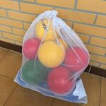 Na zdjęciu widać dużą, białą, prześwitującą siatkę stojącą na podłodze przy żółtej ceglanej ścianie. W siatce znajdują się lekkie, kolorowe piłki – głównie żółte, czerwone i zielone. Piłki są ściśnięte i wypełniają siatkę niemal do pełna. Pod siatką leży podłużny, jasny przedmiot, częściowo zasłonięty. Podłoga ma brązowe, gładkie płytki.