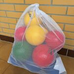 Na zdjęciu widać dużą, białą, prześwitującą siatkę stojącą na podłodze przy żółtej ceglanej ścianie. W siatce znajdują się lekkie, kolorowe piłki – głównie żółte, czerwone i zielone. Piłki są ściśnięte i wypełniają siatkę niemal do pełna. Pod siatką leży podłużny, jasny przedmiot, częściowo zasłonięty. Podłoga ma brązowe, gładkie płytki.