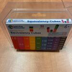 Na blacie o drewnopodobnej powierzchni stoi przezroczyste, plastikowe pudełko z kolorowym zestawem edukacyjnym. Na opakowaniu znajduje się napis „Equivalency Cubes – Fraction Tower”. W środku widoczne są pionowe klocki w różnych kolorach, ułożone obok siebie według wartości ułamków od 1.0 do małych części. W górnej części widnieją także małe ilustracje cylindrów pokazujące sposób użycia.