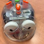 Na zdjęciu znajduje się niewielki robot o półprzezroczystej, plastikowej obudowie w kształcie kopuły. Przez obudowę widać elementy elektroniczne i przewody. Na górze znajdują się kolorowe przyciski z symbolami strzałek oraz zielony przycisk z napisem „GO”. Z przodu widoczne są duże, białe elementy przypominające oczy oraz czarna linia układająca się w symbol przypominający uśmiech. Robot stoi na gładkim, jasnobrązowym blacie.