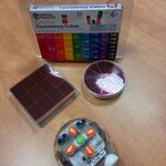 Na zdjęciu na drewnianym blacie ułożono kilka przedmiotów edukacyjnych. Na górze znajduje się pudełko z zestawem „Equivalency Cubes”, poniżej po lewej – kwadratowy element z podziałką, a po prawej – okrągły model przypominający koło podzielone na części. W dolnej części kadru widoczny jest mały przezroczysty robot z kolorowymi przyciskami.