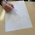 Dłoń trzymająca długopis rysuje na kartce prostą postać szkicowaną długopisem, z kilkoma krótkimi opisami zapisanymi wokół rysunku. Kartka leży na jasnym blacie.