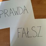 Dwie kartki leżą na drewnianej powierzchni. Na górnej widnieje napis „PRAWDA”, a na częściowo zasłoniętej dolnej — „FAŁSZ”.