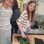 Na zdjęciu na głównym planie widać dwie dziewczynki, które patrzą się wprost do obiektywu i uśmiechają się. Jedna z nich stoi prosto, ma ręce z tyłu. Druga z nich maluje wałkiem klocki lego czarną farbą. W tle widać osobę dorosłą, która jest odwrócona tyłem oraz szafki.