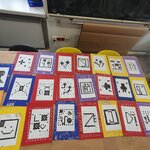 Na zdjęciu widać prace 23 uczniów, którzy wykonali je na podstawie klocków lego. Leżą one na stole. Dziewięć z nich ma czerwone tło, 6 z nich ma żółte tło, 4 mają fioletowe tło, a 4 mają granatowe tło. W tle widać parapet i tablicę kredową z wiszącym pod nią szarym papierem.