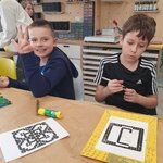 Na głównym planie widać dwójkę uczniów. Jeden z nich w granatowej bluzie z kapturem, uśmiecha się i pokazuje trzy palce, drugi z nich w czarno-białym T-shircie zamyka mazak i patrzy zamyślony w dół. Na stole przed nimi leżą dwie prace i jedna matryca z klocków lego, klej w sztyfcie. W tle widać szafki i dwie postacie.