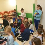 Zdjęcie na pierwszą stronę – na zdjęciu widać 20 dzieci, którzy słuchają polecenia, czytanego przez ucznia stojące i trzymającego kopertę. Czwórka uczniów stoi, dwójka klęczy, a reszta siedzi. W tle widać korytarz, jasne ściany z zielonym pasem, podjazd dla osób niepełnosprawnych oraz drzwi do pomieszczenia A-2.