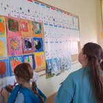 Na zdjęciu widać dzieci, które rozwiązują zagadkę. Widać dużą tablicę z alfabetem i pracami plastycznymi innych dzieci. Jedna dziewczynka z kucykiem, która stoi najbliżej tablicy zapisuje rozwiązanie zagadki, druga dziewczynka z dwoma kucykami oraz chłopiec z prawej strony zdjęcia patrzą na tablicę. W tle widać szafki dzieci , pachołek gimnastyczny i okna z zasuniętą roletą.