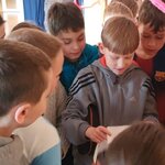 W centralnym punkcie zdjęcia widać chłopca w szarej bluzie z trzema paskami, który uśmiecha się, gdy czyta zadanie z koperty. Pozostałe osoby słuchają, przyglądają się kartce. Dziewczynka z opaską z uszami ma zaciśnięte usta. W tle widać dalszą część korytarza i przeszklone drzwi.