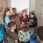 Na zdjęciu widać 10 dzieci, która stoi przed salą lekcyjną. Jedna dziewczynka czyta zadanie. Obok niej stoi dziewczynka z różową opaską z uszami zająca. W Tle widać łazienkę i białe drzwi do niej, które są otwarte.