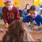  Na zdjęciu widać piątkę dziewczyn, które siedzą przy jednym stoliku i rozkładają jajka niespodzianki, które dostały od zajączka. Trzy z nich: z opaską różowymi uszkami, w niebieskiej bluzie i kremowym sweterku uśmiechają się. Jedna z nich jest poważna i skupiona na złożeniu zabawki. Jednej dziewczynki twarzy nie widać, gdyż jest odwrócona tyłem. W tle widać dziewczynkę w szaro-niebieskiej bluzie, która je czekoladkę oraz szafki dzieci i ich plecaki.
